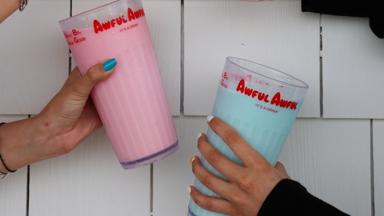 Maincured hands holding Awful Awful cups containing milkshakes, one pink and one light blue.
