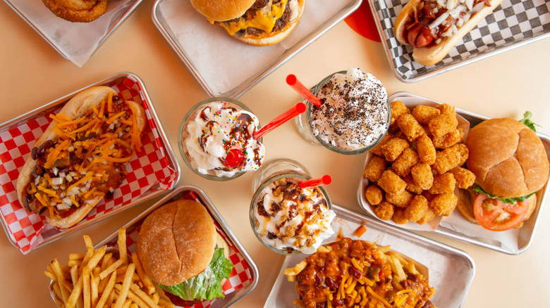 Three milkshakes with red straws and toppings, surrounded by trays of food.
