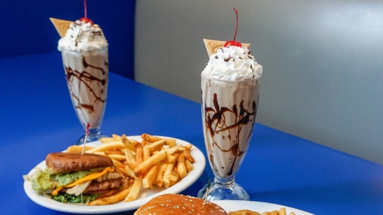 Two milkshakes in old-fashioned glasses, on blue table with plate of burger and fries.
