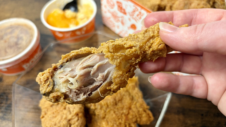 Hand holding a Popeyes Signature chicken drumstick with more fried chicken and side dishes in the background