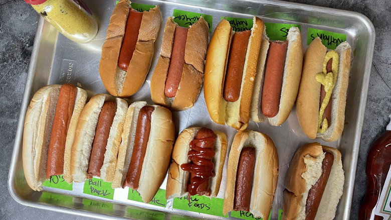A tray of 11 different types of hot dogs with several bites out of each hot dog.