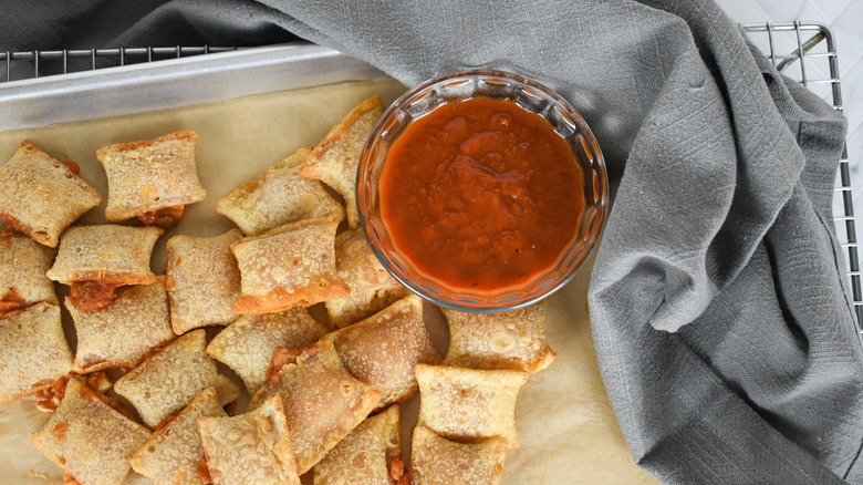 Pizza rolls on a parchment-lined sheet tray with a bowl of marinara sauce and a grey towel