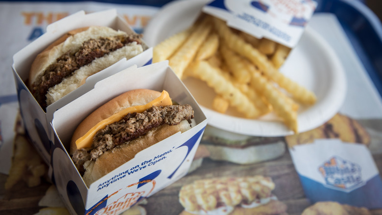 Two small White Castle slider hamburgers in cardboard boxes with french fries on a paper plate