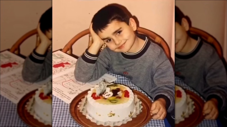 a photo of a young CZN Burak in a gray sweater smiling at the camera, a cake and open book on the table before him