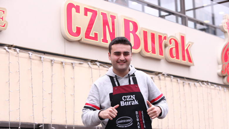 CZN Burak, also known as Burak Ozdemir, poses with his thumbs up and smiles brightly in front of his restaurant's signage