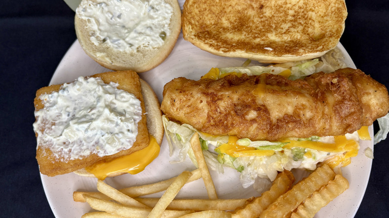 Two open fish sandwiches with french fries on a white plate on a dark background