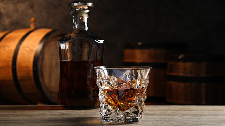 A beveled glass of bourbon on a wood table in front of a glass bottle and oak barrels