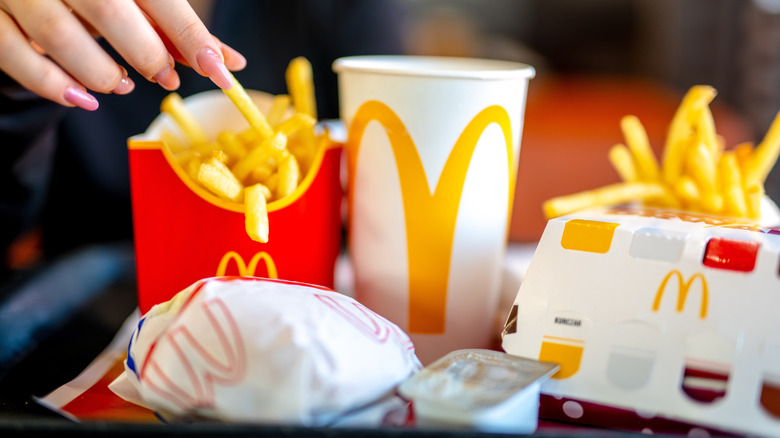 A hand picks up a fry from a tray with McDonald's french fries, burgers, drink, and dipping sauce