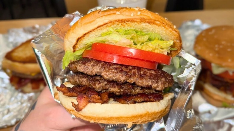 Hand holding Five Guys hamburger with two patties, bacon, lettuce, and tomato.