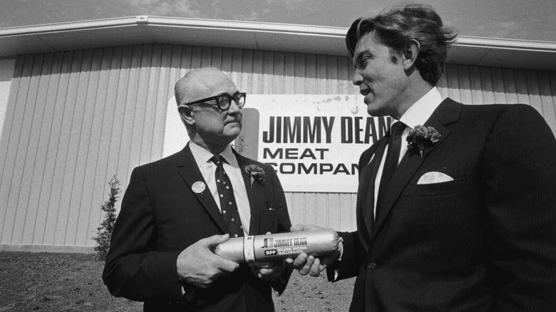 Jimmy Dean and a man hold a Jimmy Dean sausage while standing outside a building of the Jimmy Dean Meat Company