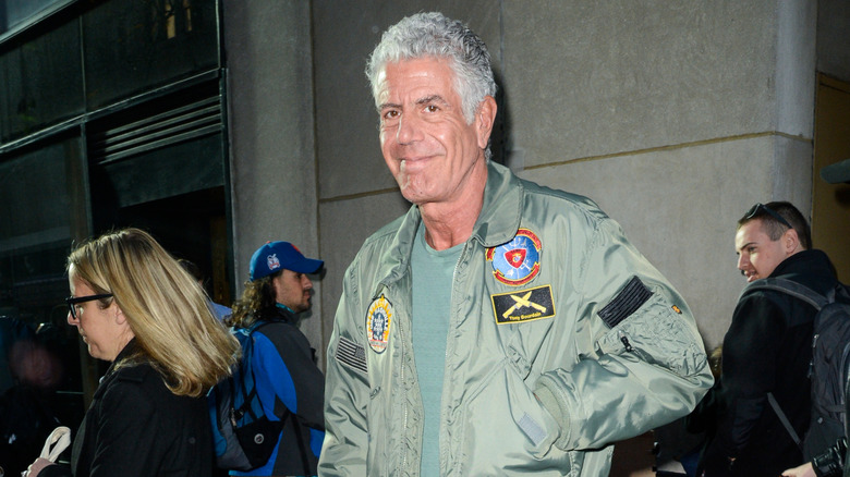 Anthony Bourdain standing in a bustling city street, smiling with one hand in his jacket pocket