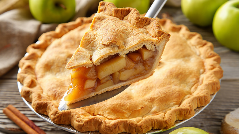 Slice of apple pie being removed from the full pie with apples surrounding it