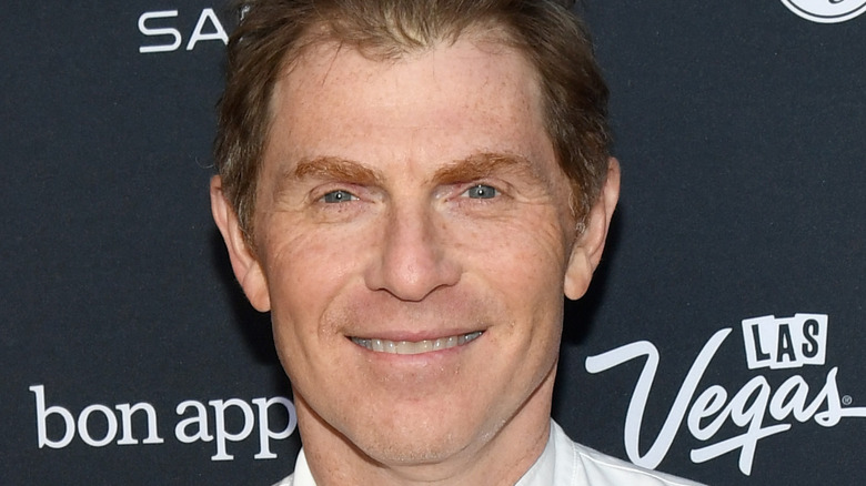 Bobby Flay wearing a chef's shirt, standing against a black and white step and repeat