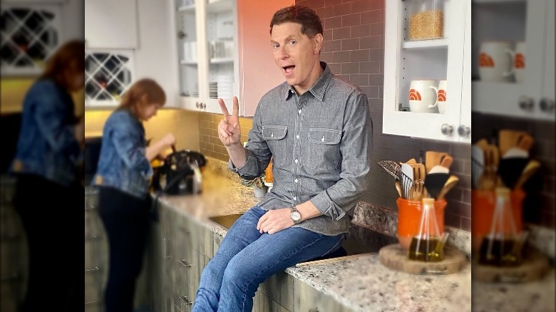 Bobby Flay sitting on a flat cooktop while showing a peace sign.