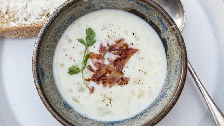 Bowl of clam chowder on white plate with metal spoon and slice of bread with butter with bacon & herb