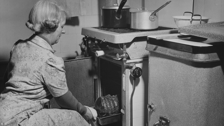 Middle-aged home cook checking a roast in front of an old-fashioned stove in a black and white photo.