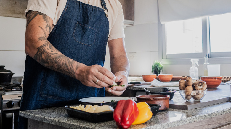 Older, tattooed home cook in blue apron and white shirt preparing raw meat, peppers, and mushrooms.