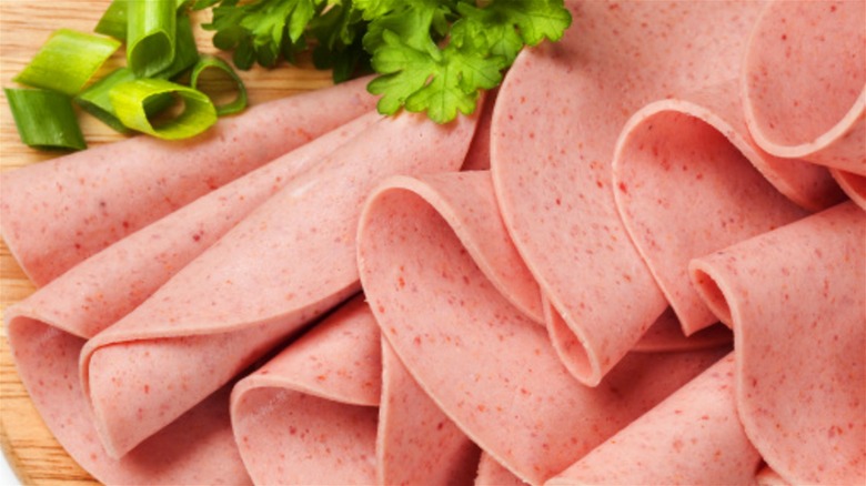 Slices of bologna meat on a wooden board with fresh greens.