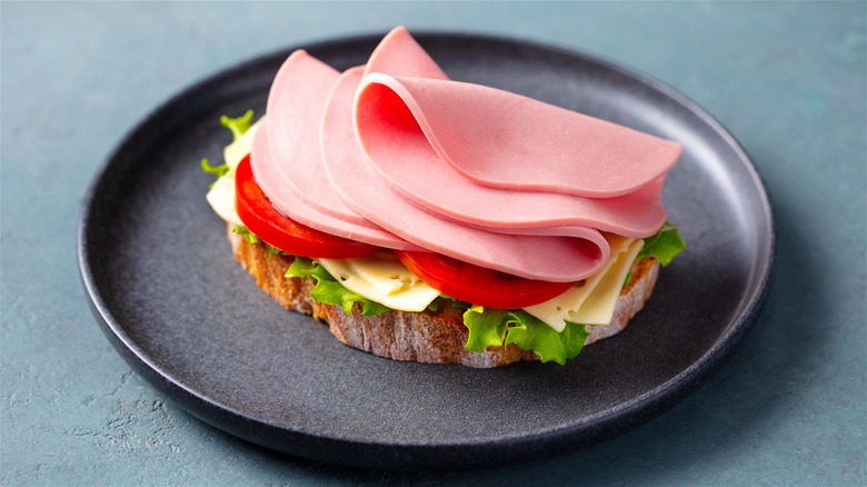 An open-faced deli sandwich with bologna, tomatoes, cheese, and lettuce on a black plate.