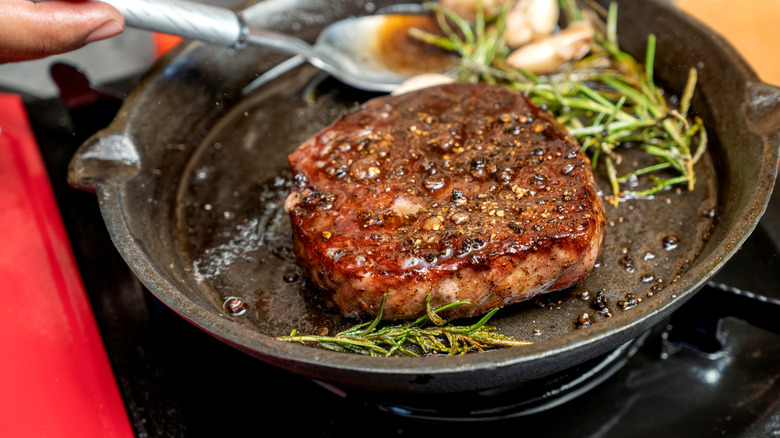A steak is cooked in a cast iron pan with herbs and garlic on a stovetop