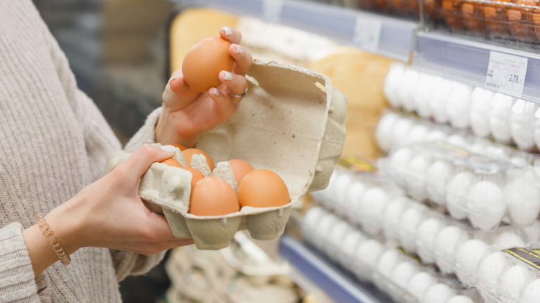 Hands holding open brown egg carton in front of eggs in the refrigerated egg section at a grocery store