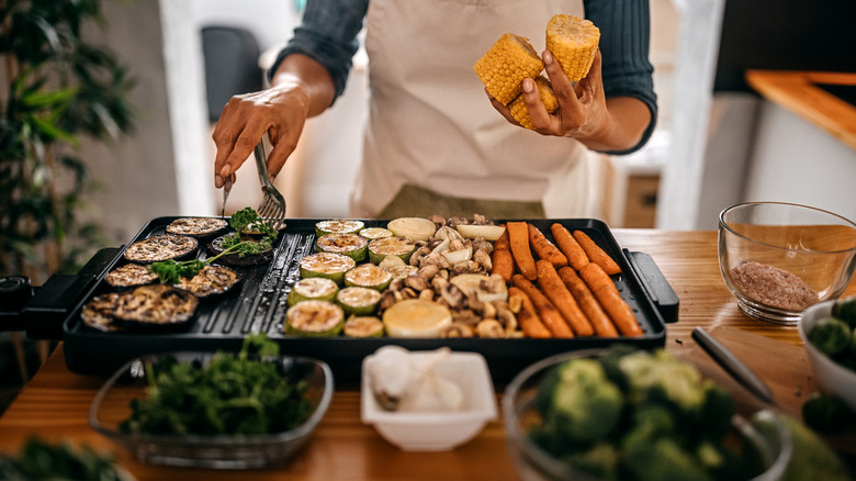 A woman in an apron holding corn on the cob and using tongs to flip grilled vegetables