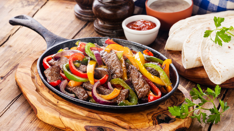 Beef fajitas in a cast-iron pan next to flour tortillas, cilantro, and condiments