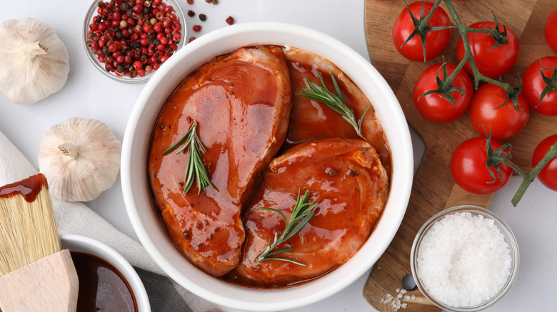 Meat marinating with fresh herbs next to a basting brush, heads of garlic, tomatoes on the vine, salt, and peppercorns