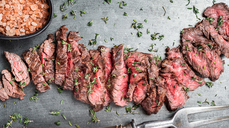Sliced skirt steak garnished with fresh herbs next to a bowl of Himalayan salt and a carving fork
