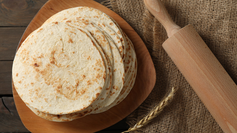 Homemade flour tortillas stacked on a wooden plate next to a rolling pin and wheat