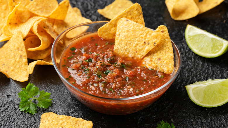 A glass bowl filled with homemade salsa garnished with fresh herbs and lime wedges surrounded by tortilla chips