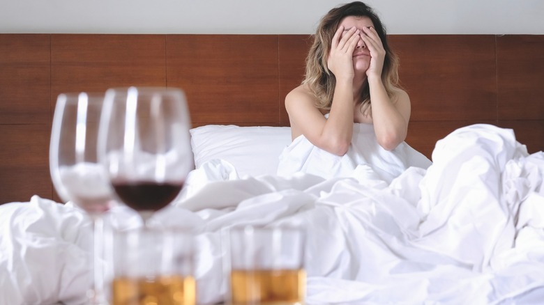 Person squeezing eyes shut with hands in bed with wine glasses in foreground