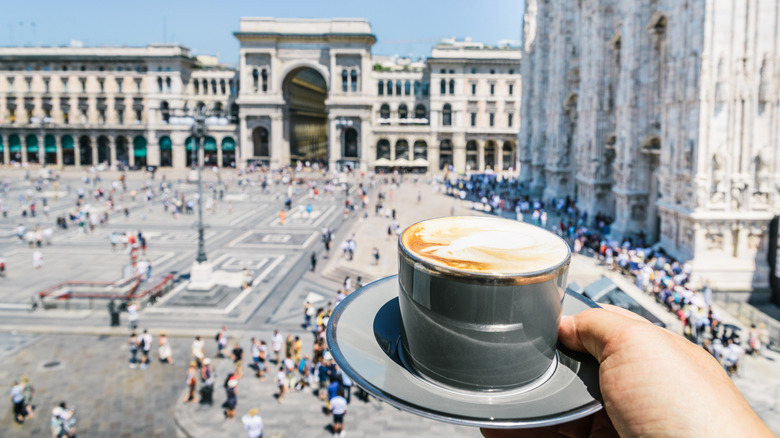 Hand holding full coffee cup on saucer with Italian architecture and crowds in background.