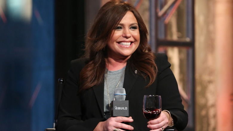Rachael Ray smiling on stage holding a microphone and a glass of red wine