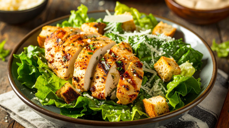 Salad topped with grilled chicken, croutons, and grated cheese over romaine lettuce on a crowded table.
