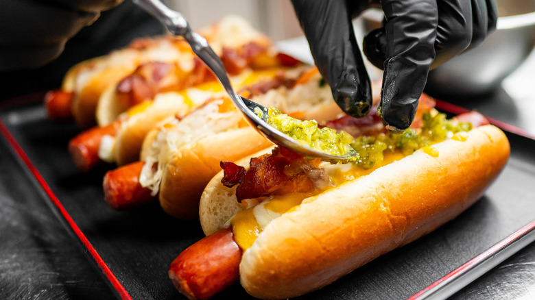 Gloved hand adding relish to row of four hot dogs on tray