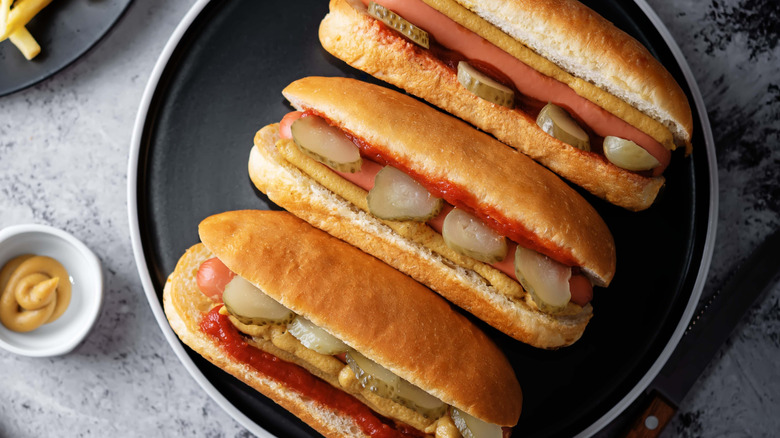 Three hot dogs topped with ketchup, mustard, and pickles on black plate