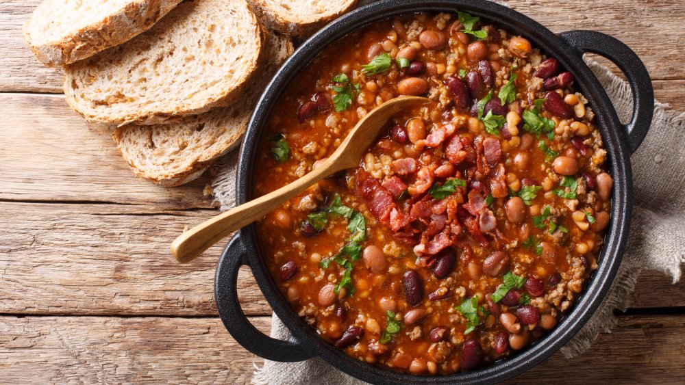 A pot of crock Crock Pot with beans and bacon inside and a serving of bread for dipping besides it. 