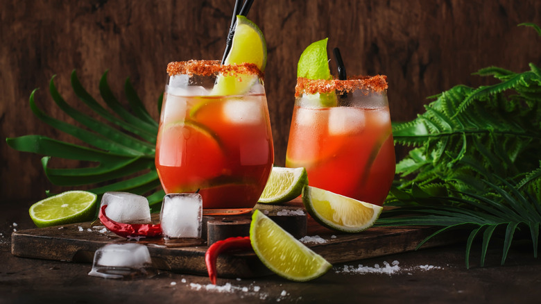 Two micheladas surrounded by lime slices, ice cubes, and dried chili peppers