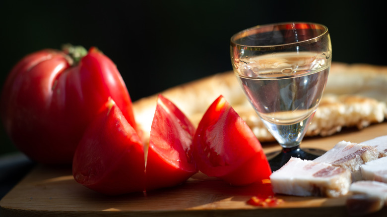 Closeup of sliced tomato, shot of vodka, and cured meat on board