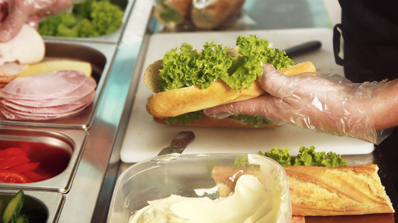 Person building a submarine sandwich with ham, tomatoes, cheese, and lettuce.