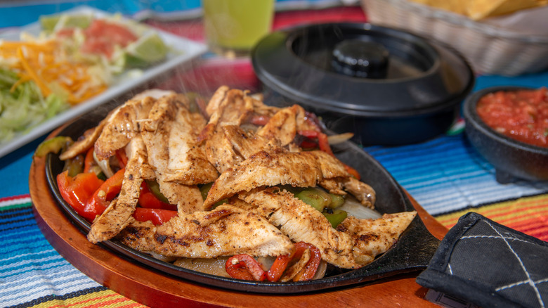 Sizzling chicken strips and bell peppers in individual hot plate on table surrounded by fajita fixings.