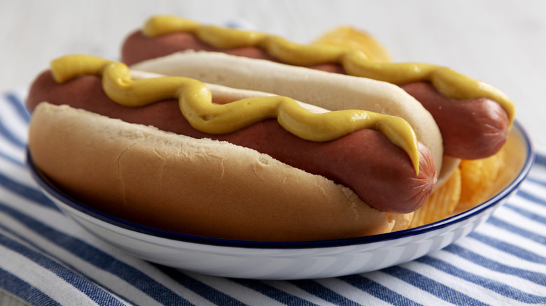 Two hot dogs with mustard in buns on blue-rimmed white plate atop blue-and-white striped towel.