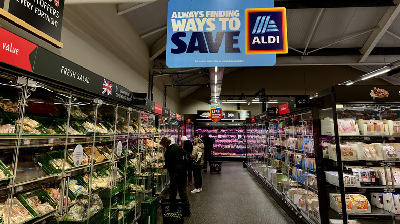 An Aldi "always finding ways to save" sign hangs over an aisle of refrigerated cases.
