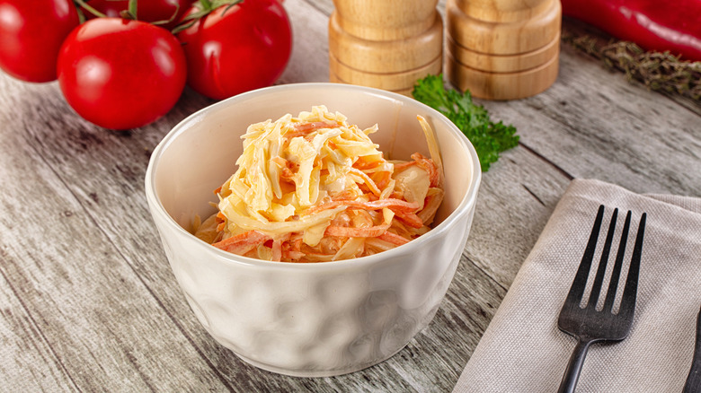 A small bowl of coleslaw on a wooden table with fork and tomatoes