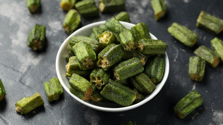 Cut okra in white bowl atop dark surface with more cut-up okra pieces on it