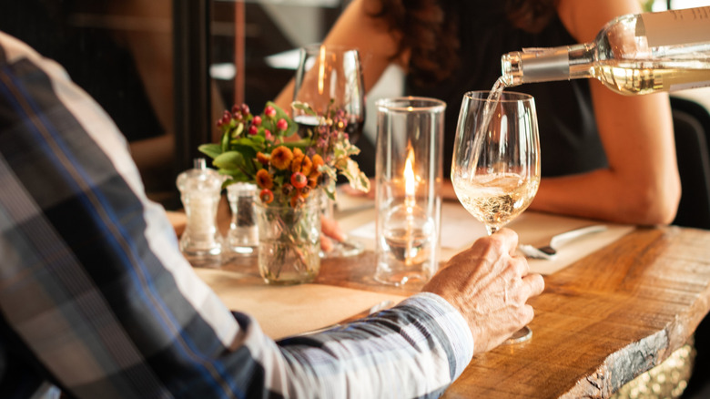A man and a woman sitting at a wooden table in a restaurant with a waiter pouring wine