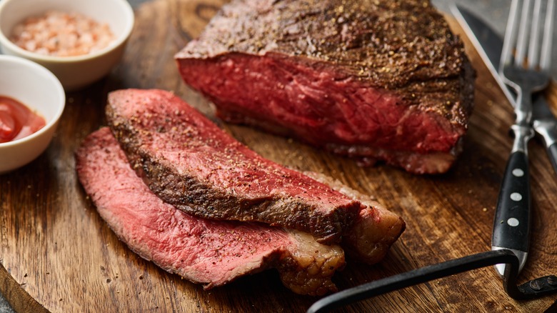 Sliced steak on wooden platter with silverware and dips 