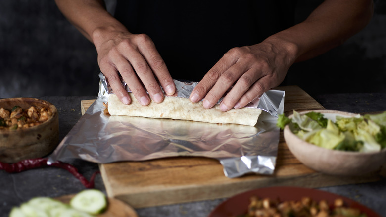 Person's hands preparing a wrap inside aluminum foil on a cutting board with green vegetables in bowls