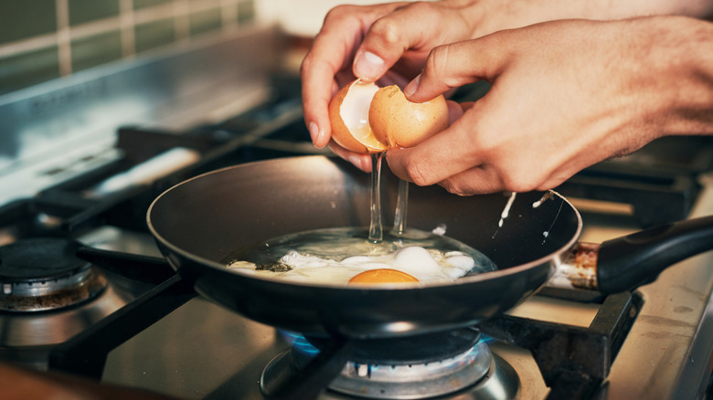Hands cracking egg into pan on stove.